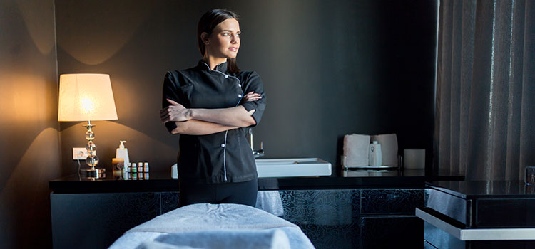 massage therapist stands in front of credenza with oils and by massage bed