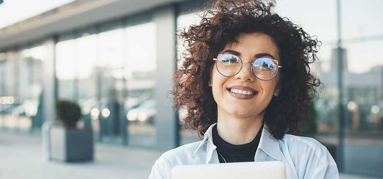 female student in glasses holds laptop outside glassfront building