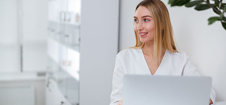 esthetician looking up licensing requirements on laptop in white on white spa