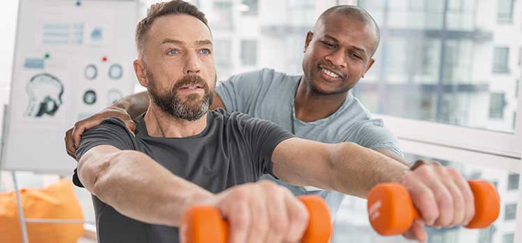 physical therapist corrects patient stance while he holds weights in front of him